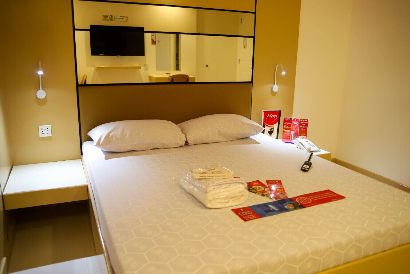 Habitación de hotel con cama, televisor de pantalla plana montado encima, lámparas de noche, teléfono y cama hecha con cuidado, incluyendo una toalla doblada y una revista.