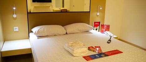 Habitación de hotel con cama, televisor de pantalla plana montado encima, lámparas de noche, teléfono y cama hecha con cuidado, incluyendo una toalla doblada y una revista.