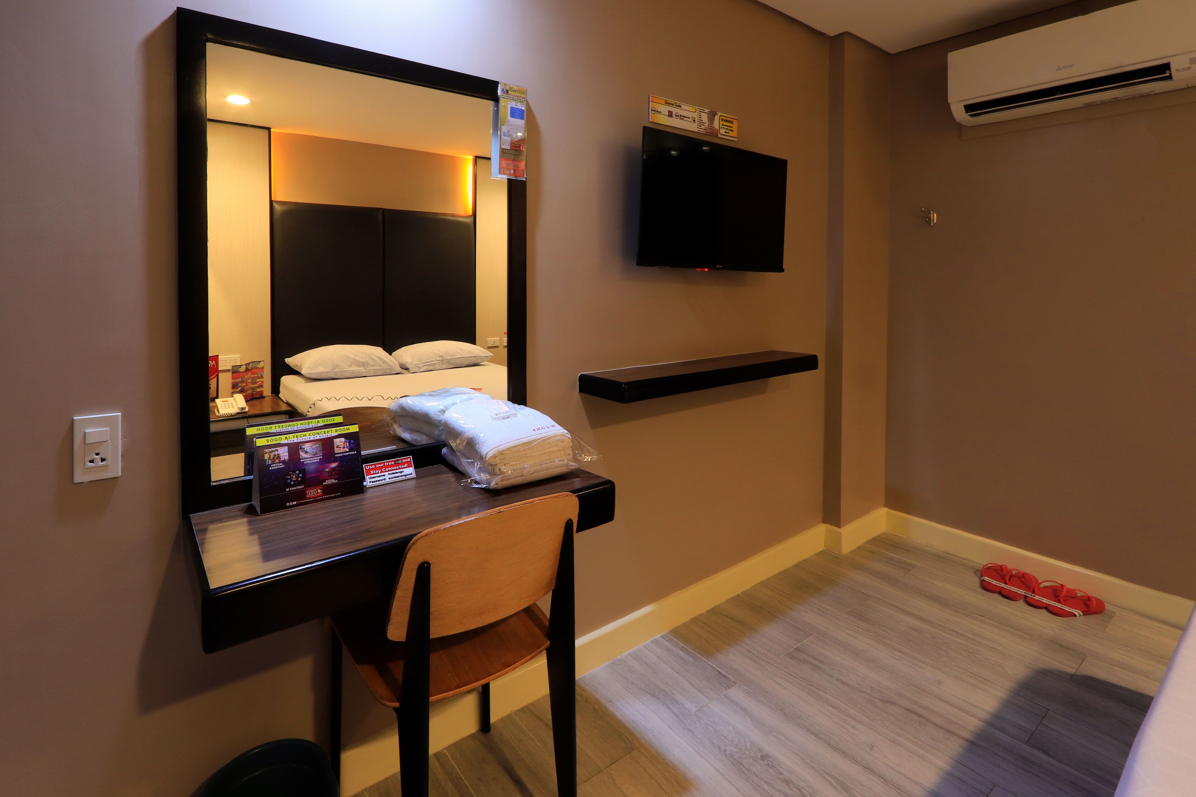 A hotel room with a bed, a desk with a chair, a TV mounted on the wall, and an air conditioning unit.
