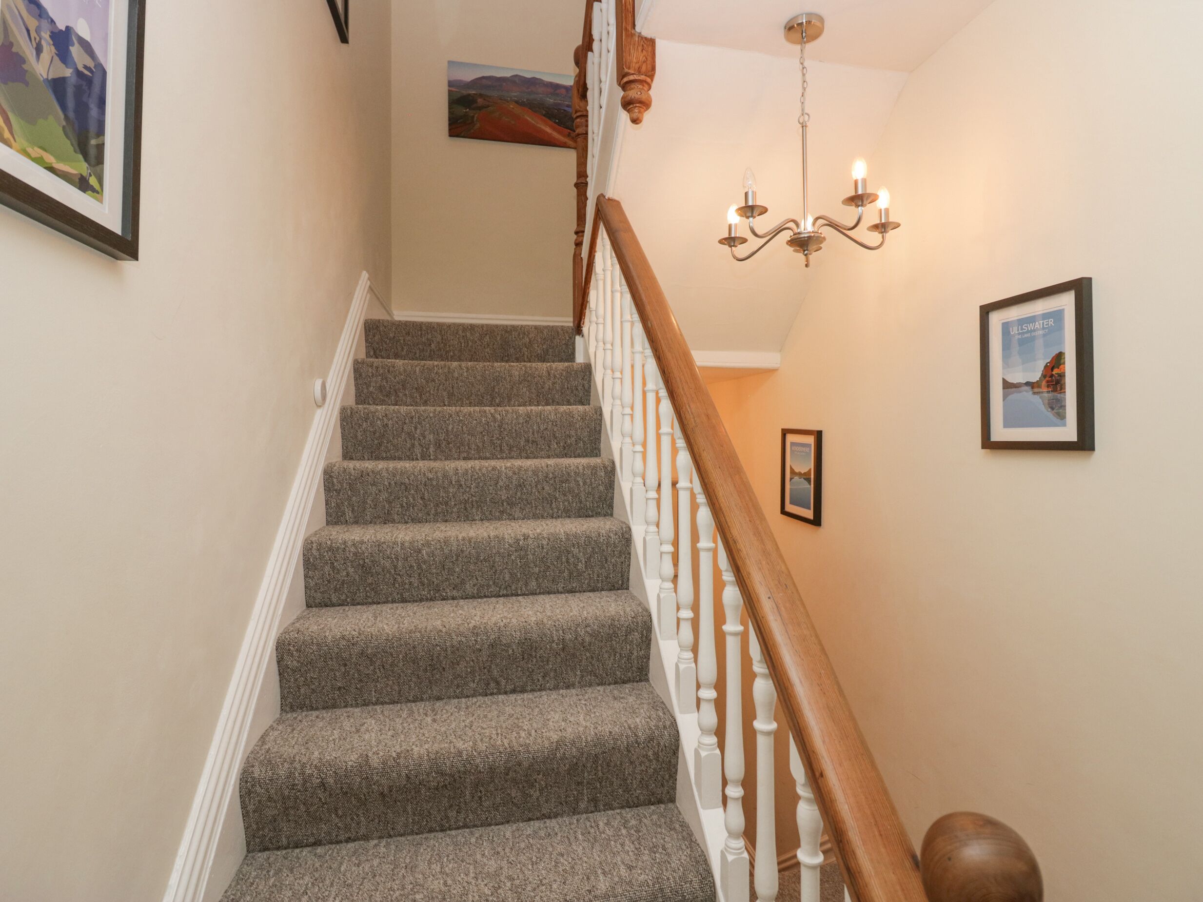 A carpeted staircase with white balusters, a chandelier, and framed pictures on the walls.