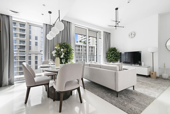 A modern living room with a dining area, large windows, and a flat-screen TV. - StoneTree - Luxurious 1 BR with Private Beach Access (Dubai)
