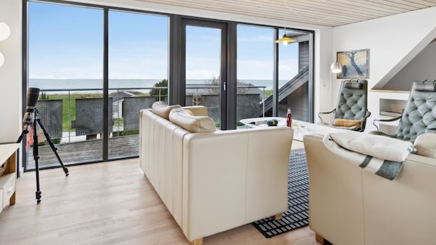 Ein modernes Wohnzimmer mit einem großen Schiebeglastür, die auf einen Balkon führt, ein weißes Sofa, ein kleiner Tisch mit einer Flasche und ein Teleskop auf einem Stativ.