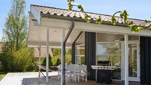 Een overdekt terras buiten met een barbecue, witte stoelen en een eettafel.