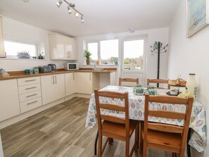 A kitchen with wooden cabinets, a microwave, and a dining table with chairs. - The Bell Apartment (Cemaes Bay)
