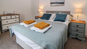 A neatly made bed with folded towels, a bedside table with a lamp, a dresser with drawers, and a painting on the wall.
