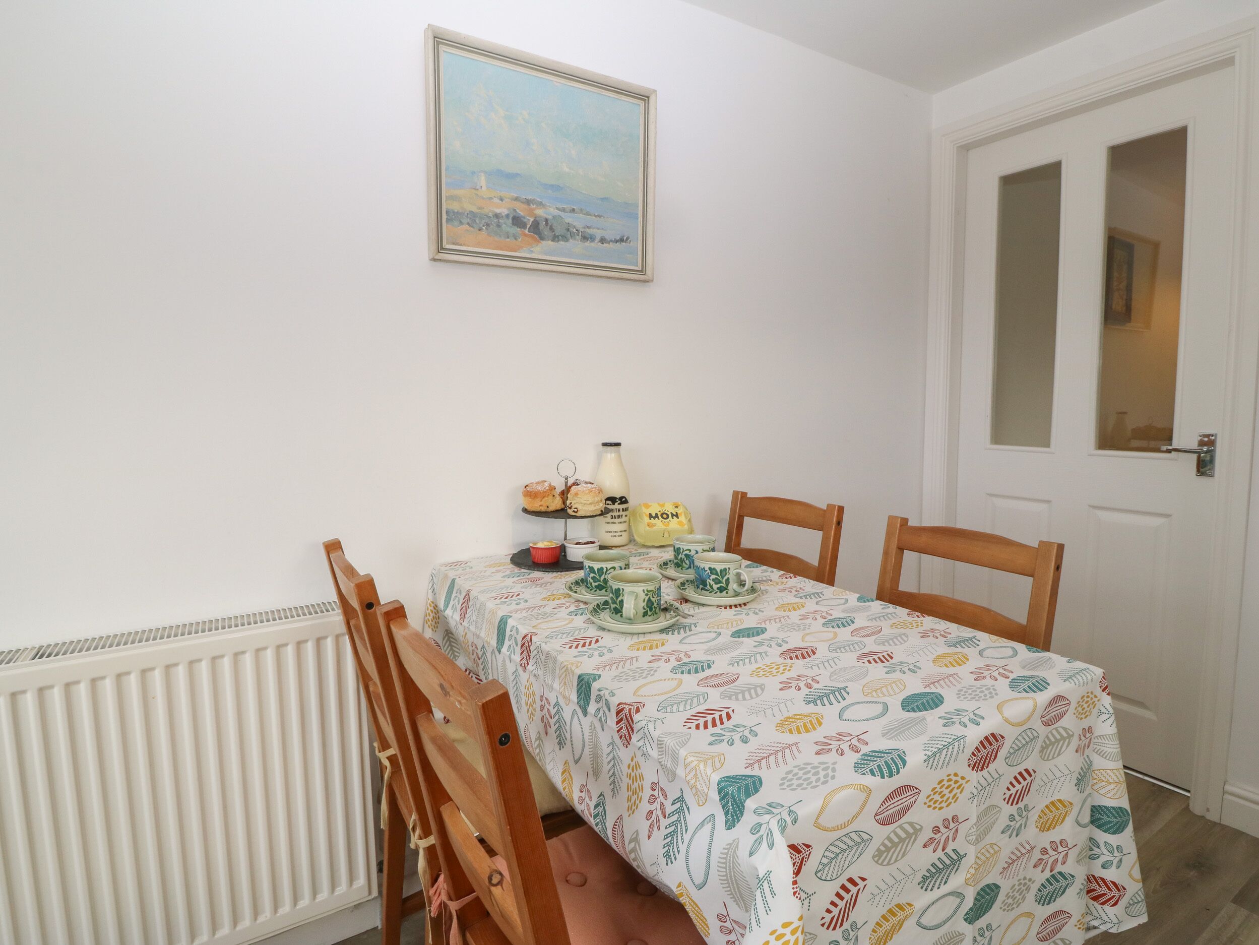 A dining area with a table set for two, featuring a patterned tablecloth, a painting above the table, and a radiator below.