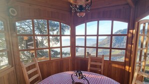 Innenansicht einer Holzhütte mit einem runden Tisch, bedeckt mit einer rot-weißen Karotischdecke, einer Hängelampe und großen Fenstern mit Blick ins Freie.