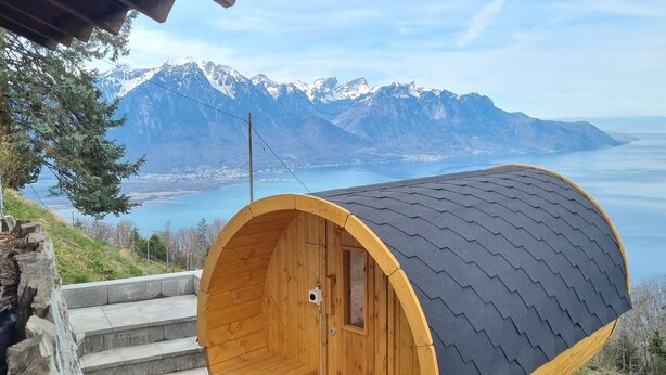 Eine hölzerne Sauna mit gewölbtem Dach, eingebettet in einen Hügel mit Blick auf Berge und einen See.