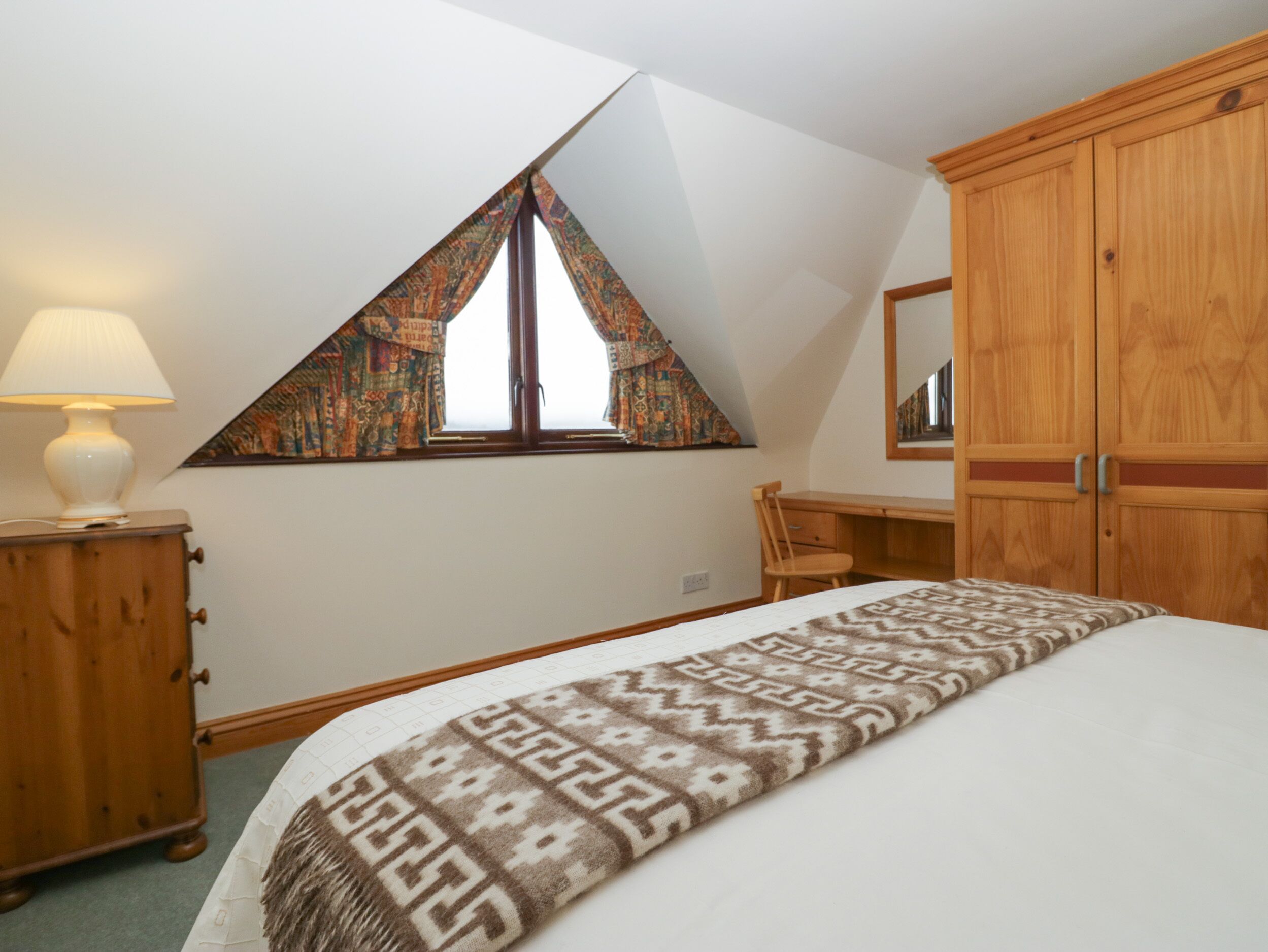 Une chambre avec un plafond en pente, un lit recouvert d’une couverture à motifs, un bureau en bois, une chaise, une lampe et une fenêtre ornée de rideaux à motifs.