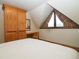 A bedroom with a sloped ceiling, a wooden wardrobe, a desk, and a window with patterned curtains.