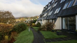 Une rangée de maisons modernes, dotées de toits inclinés et de grandes fenêtres, est entourée de jardins bien entretenus et un chemin dégagé mène à un bâtiment traditionnel situé au fond.