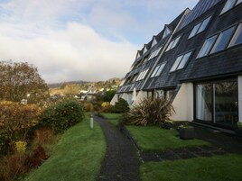 A row of modern houses with sloped roofs and large windows, surrounded by well-maintained gardens and a clear path leading to a traditional building in the background.