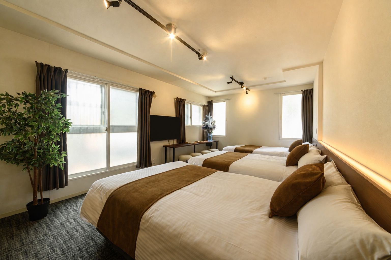 A hotel room with three beds, a flat-screen TV, a potted plant, and a large window.