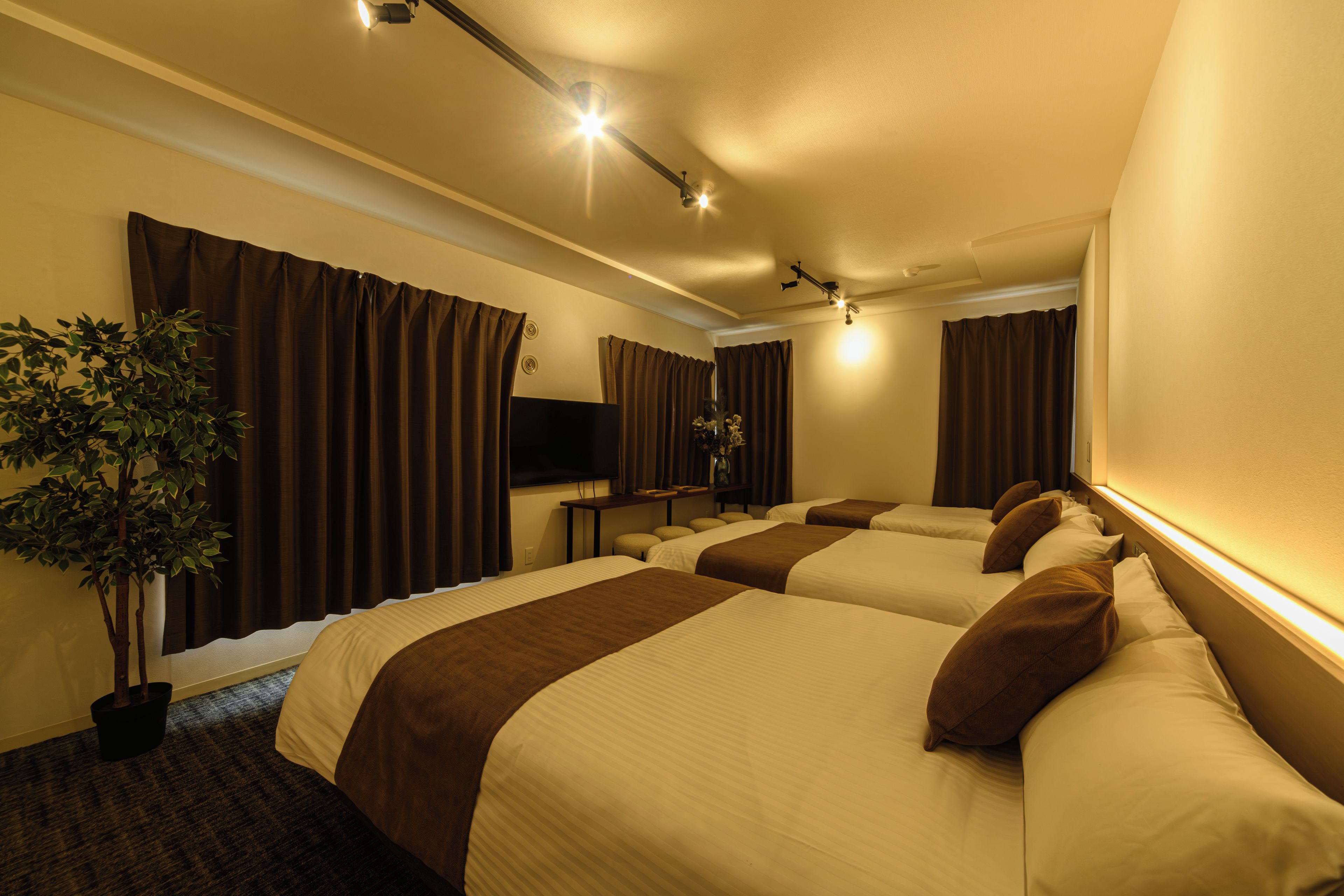 Een hotelkamer met vier bedden, een flatscreen televisie, een kamerplant en een groot raam met gordijnen.