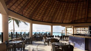 An outdoor dining area with tables and chairs, a thatched roof, and a view of the ocean. - Westin Lagunamar Ocean Resort - Luxurious 1-Bedroom Villa - Family Friendly Resort! (Cancun)