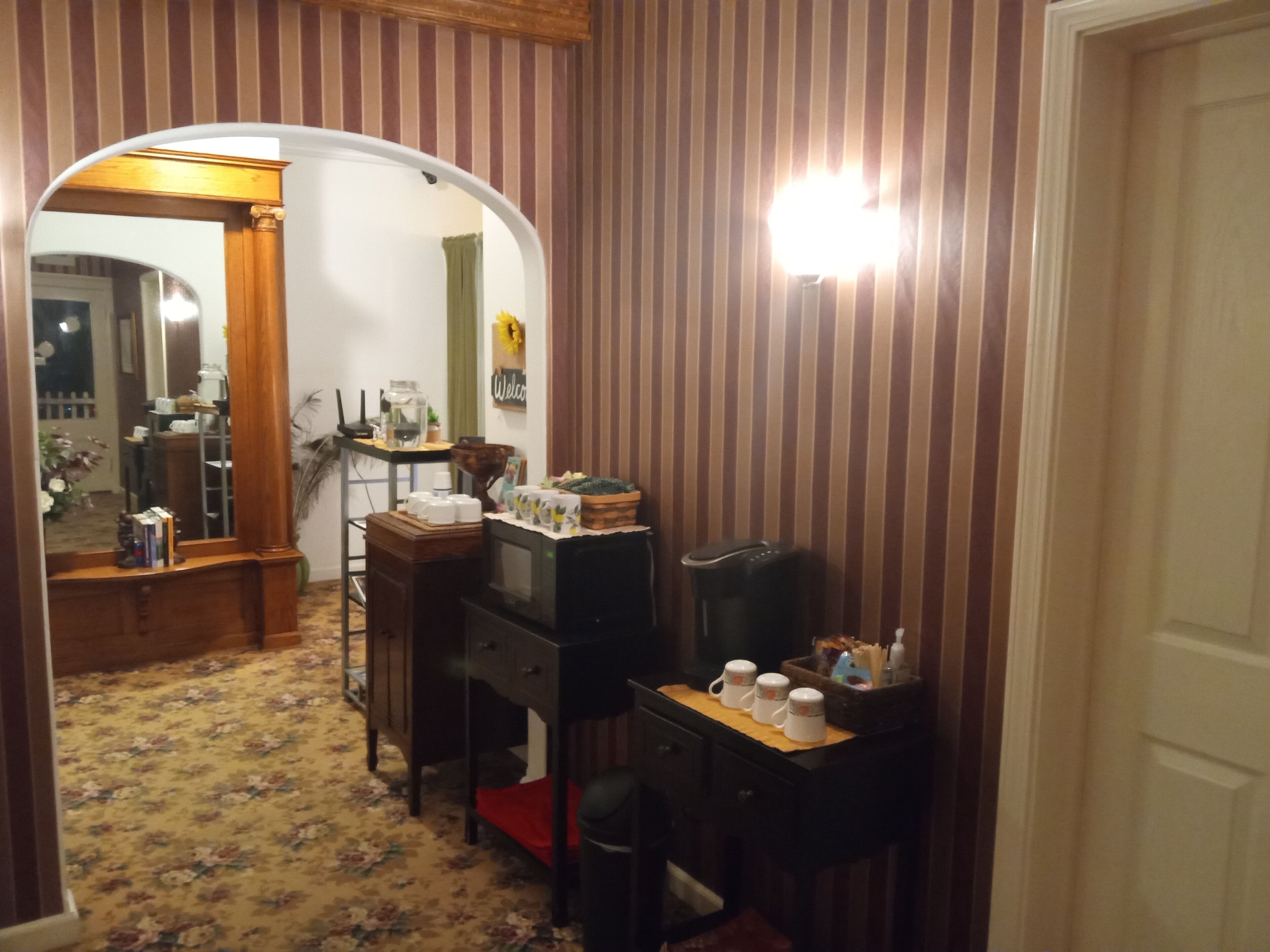 Une pièce avec un papier peint à rayures, un bureau en bois, une chaise, une petite table avec des tasses et un miroir qui reflète une porte et une plante.