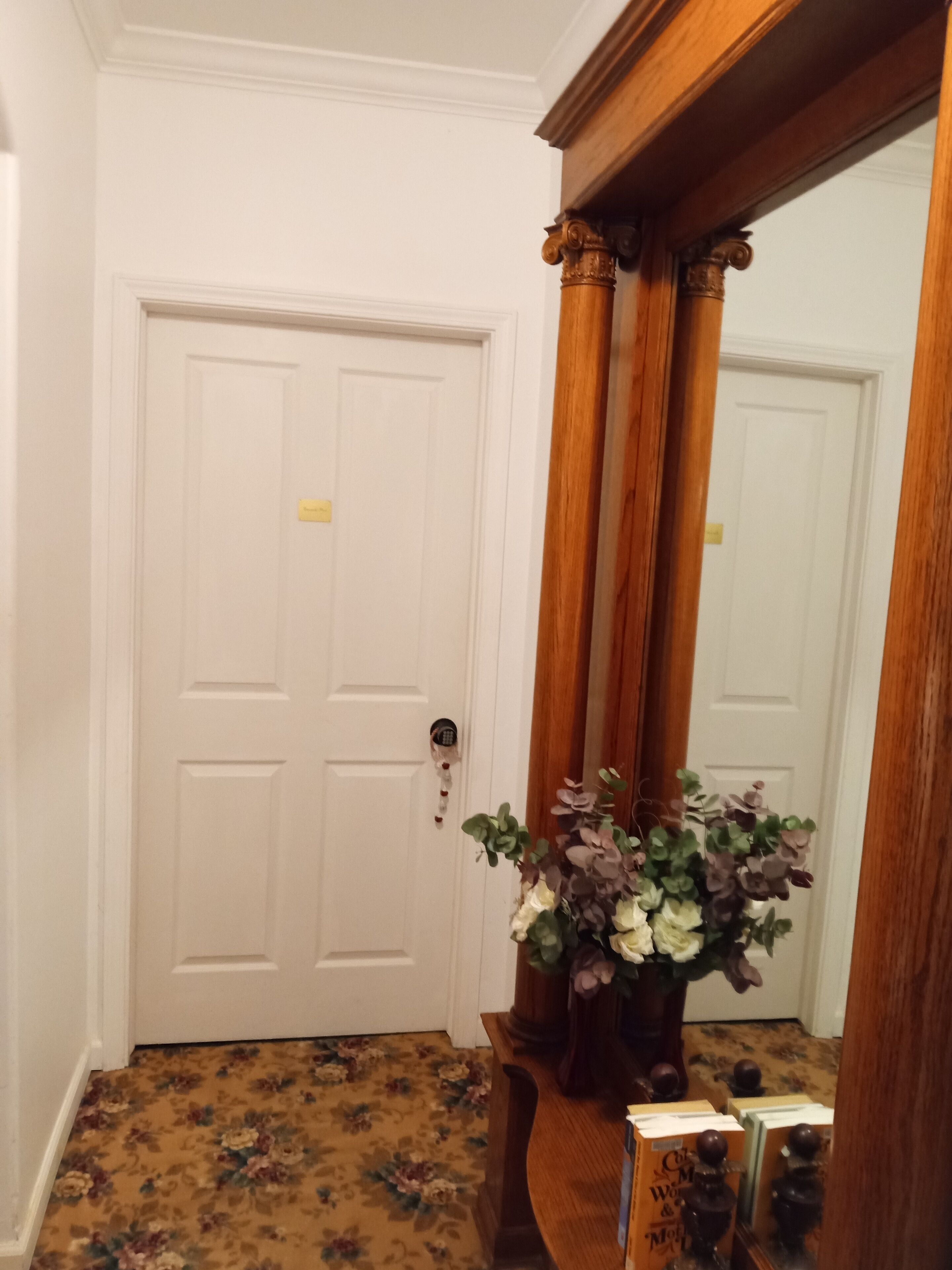 Um corredor com uma porta branca, um espelho de madeira e um tapete com padrão floral.