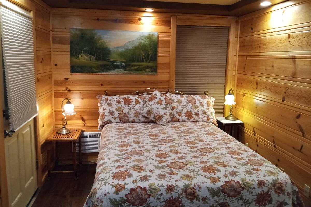 A wooden-paneled bedroom with a bed, two bedside lamps, a painting on the wall, and a window with blinds.