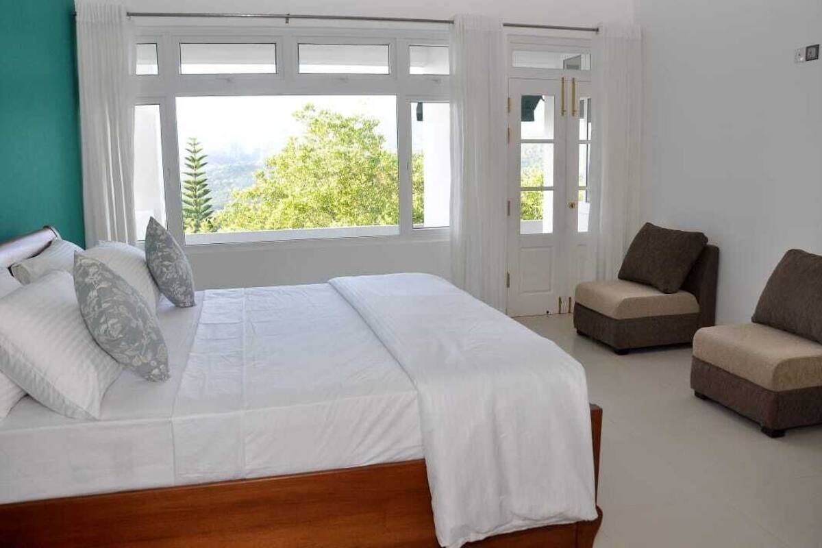 A bedroom with a large bed, two armchairs, and a view of greenery through the window.