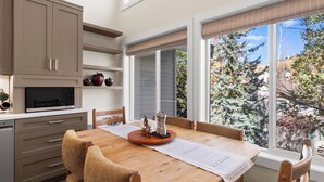 Cocina moderna con comedor, mesa de madera y ventanas amplias que dan a vista de árboles y una casa.