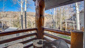 Una terraza de madera con una silla de mimbre, con vistas a un paisaje montañoso de árboles desprovistos de hojas y un cielo azul despejado.