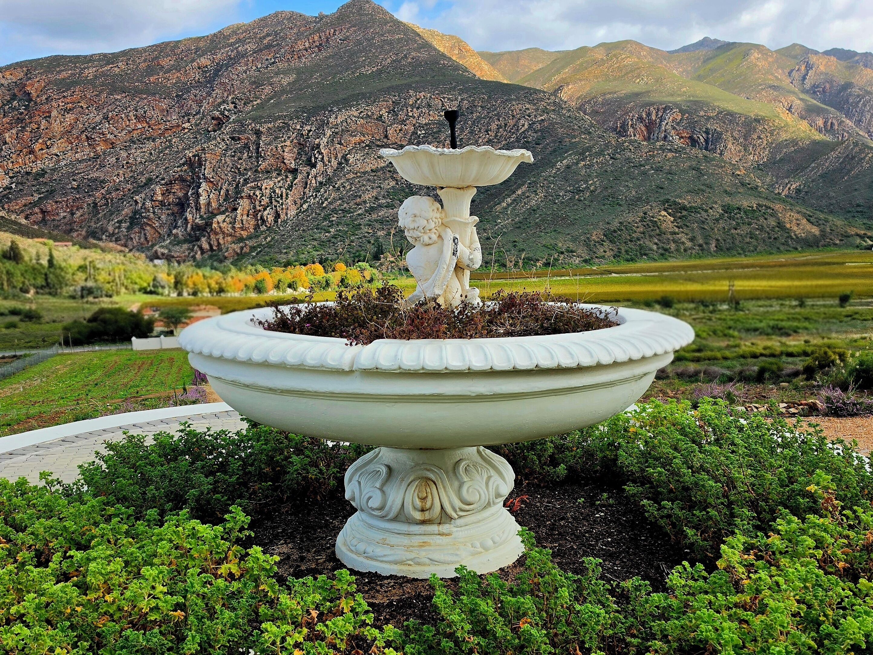 A white outdoor fountain with a statue in the center, surrounded by greenery, with mountains in the background.