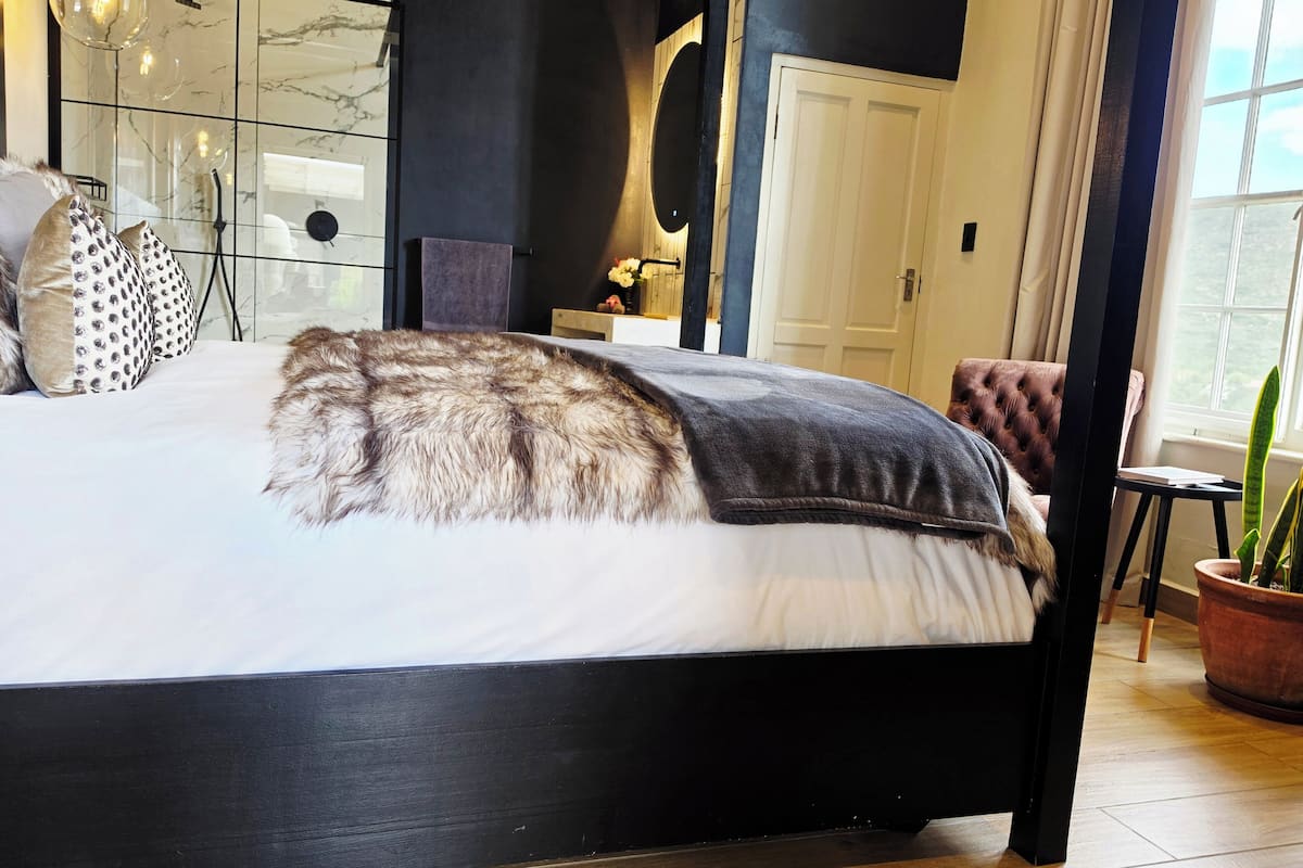 A bedroom with a bed, a nightstand, a chair, a potted plant, and a bathroom with a glass door.