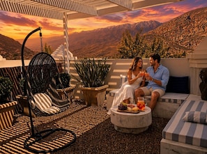 Una pareja sentada en un balcón con vistas a montañas, una silla colgante y una mesa con aperitivos.