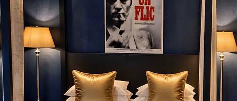 A hotel room with a large poster on the wall, two bedside lamps, and a bed with two gold pillows.
