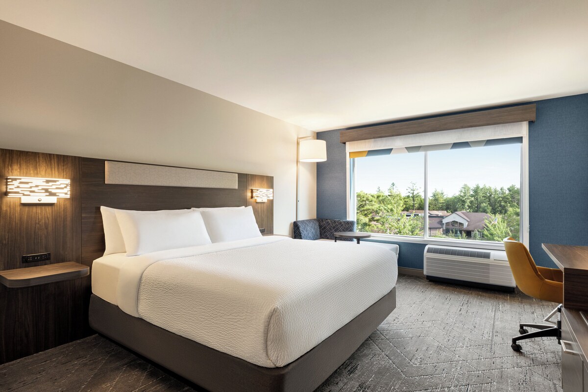 A modern hotel room with a large bed, a desk, and a chair. There is a window with a view of trees and houses.