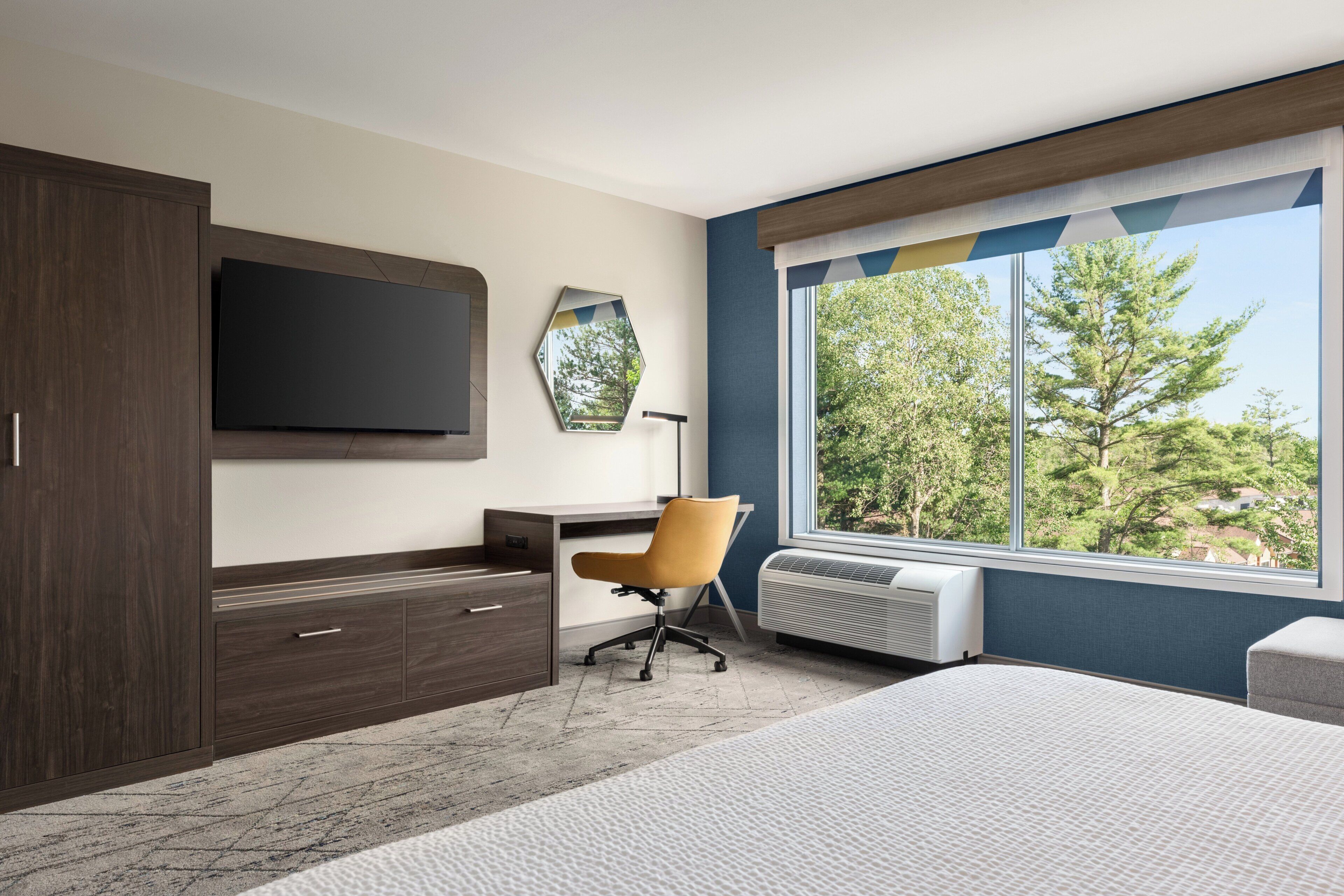 A modern hotel room with a large bed, a desk with a chair, a flat-screen TV, a mirror, and a window with a view of trees.
