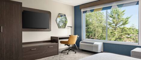 A modern hotel room with a large bed, a desk with a chair, a flat-screen TV, a mirror, and a window with a view of trees.