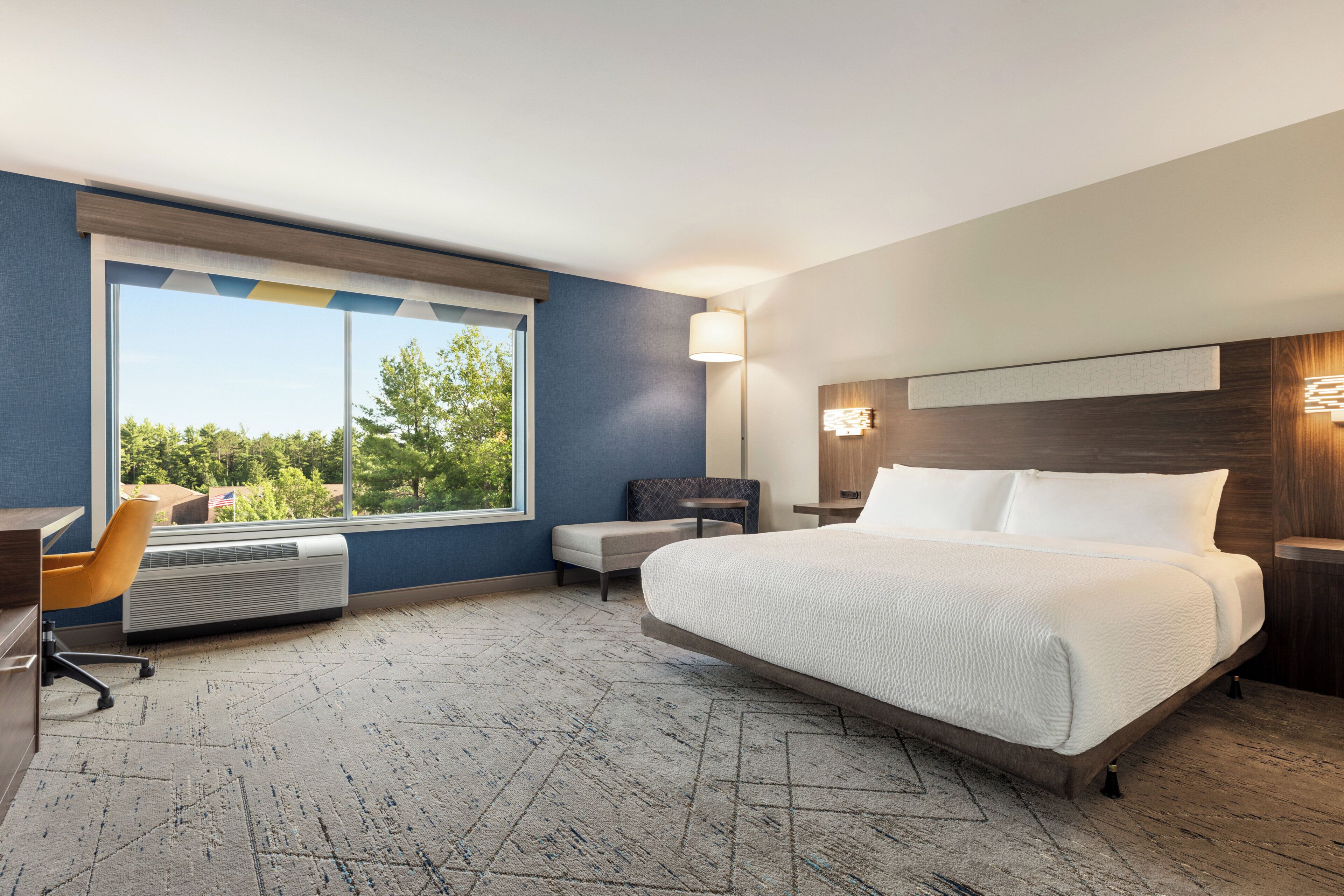 A hotel room with a large bed, a desk, a chair, and a window with a view of trees.