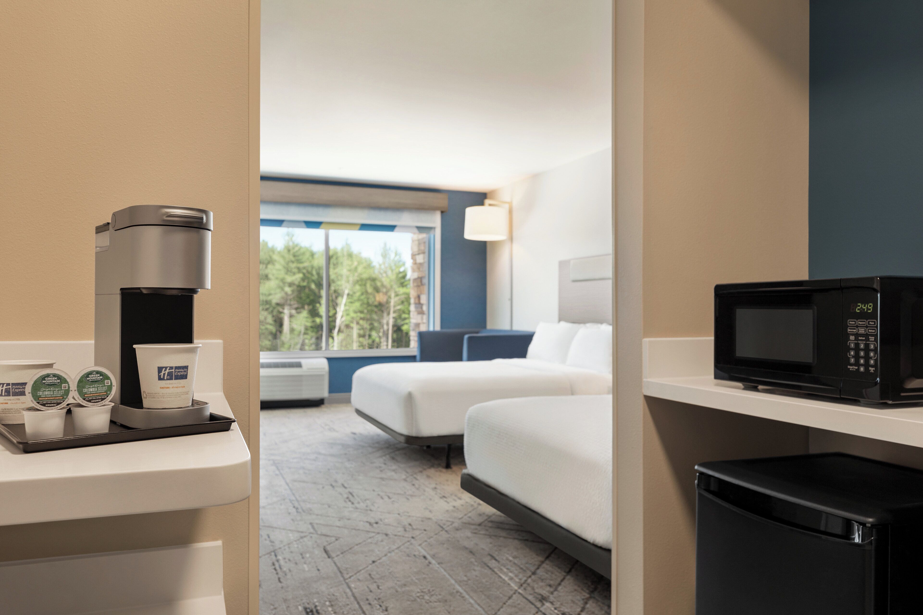 Ein Hotelzimmer mit Kaffeemaschine, Mikrowelle und einem kleinen Tisch mit Tassen. Es gibt zwei Betten und ein Fenster mit Blick auf Bäume.