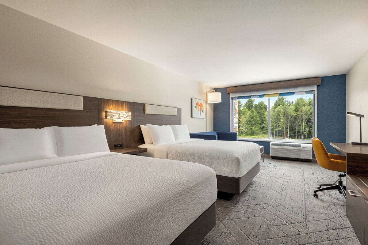 A hotel room with two beds, a desk, a chair, a window with a view of trees, and a wall-mounted lamp.