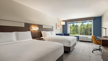 Habitación de hotel con dos camas, un escritorio, una silla, un ventanal con vistas a árboles y una lámpara de pared.