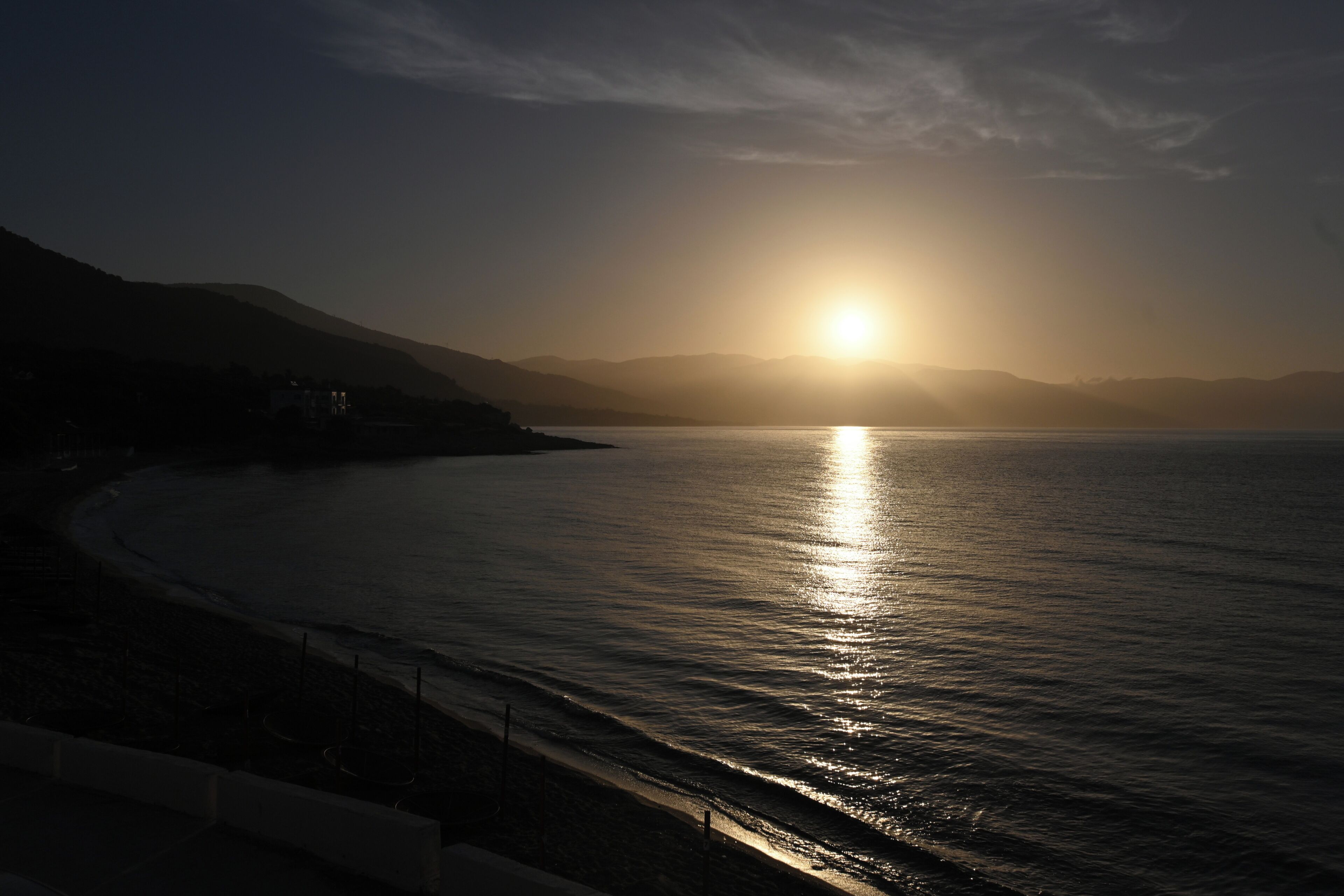 Un coucher de soleil sur la côte, avec une mer calme, une plage et des montagnes à l’horizon.