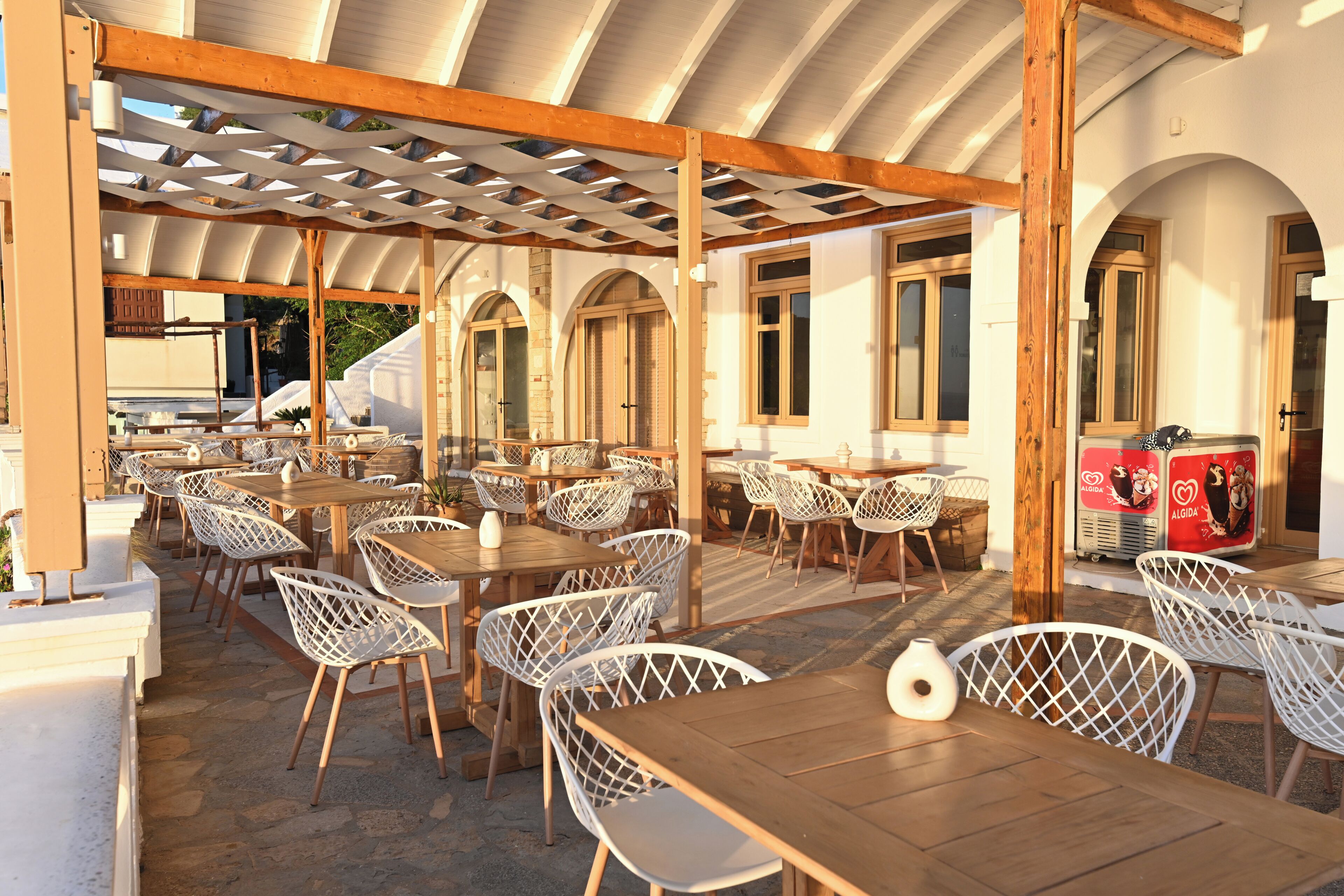 An outdoor café with wooden tables and white lattice chairs, under a wooden pergola with a curved roof.
