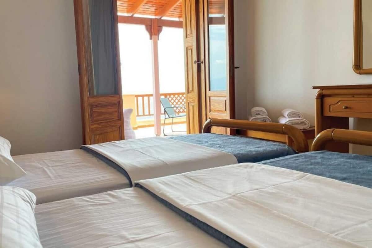 A room with three beds, a wooden desk, and a balcony with chairs.