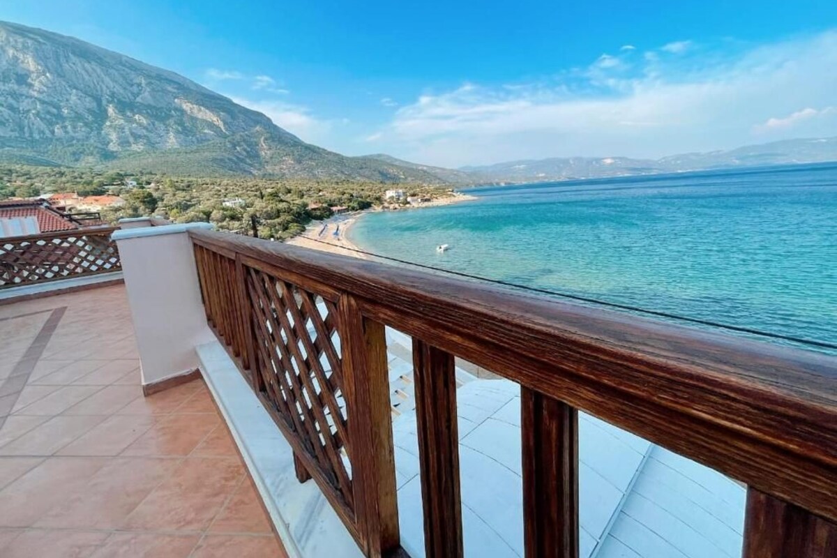 Un balcón con barandilla de madera con vistas a una zona costera de aguas azules cristalinas y montañas al fondo.