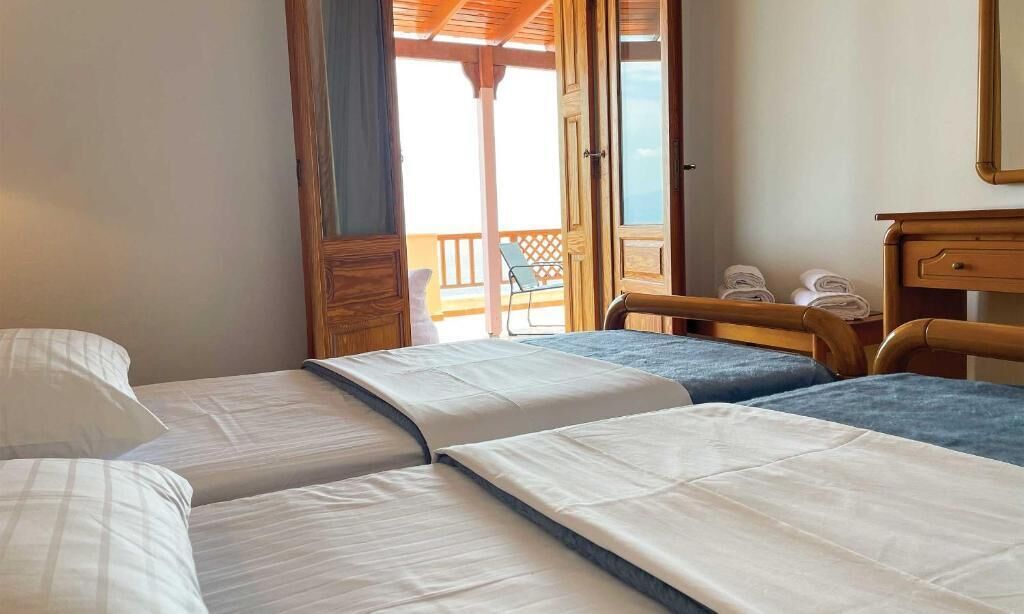 A room with three beds, a wooden desk, and a balcony with chairs.