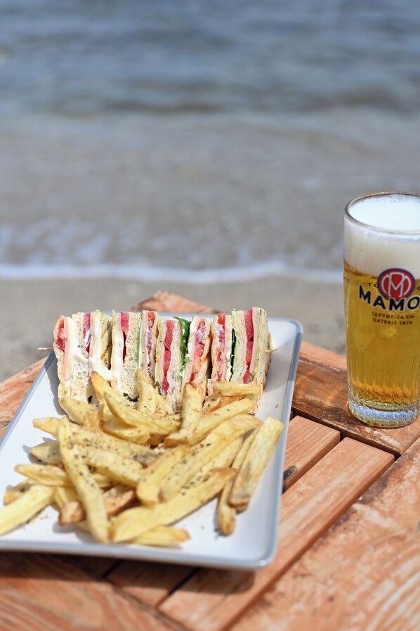 A plate of sandwiches and fries on a wooden table with a glass of beer. - Nereides Seaside Apartments (Samos)