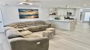Un salon moderne avec un canapé d’angle, un coin cuisine et une grande fenêtre donnant sur un paysage au coucher du soleil.