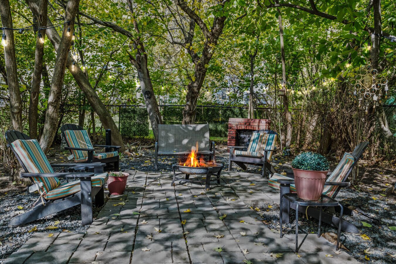 An outdoor seating area with chairs and a fire pit, surrounded by trees and a brick fireplace.