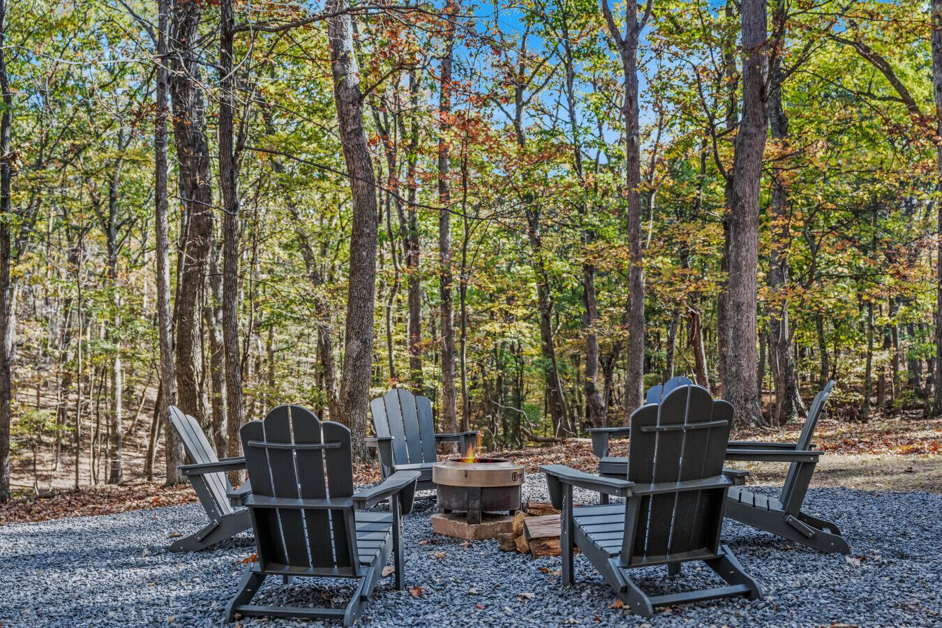 Un décor forestier avec quatre chaises Adirondack disposées autour d’un feu de camp.