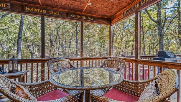 A covered outdoor seating area with wicker chairs and a glass table, surrounded by wooden railings and large windows offering a view of a forested area.