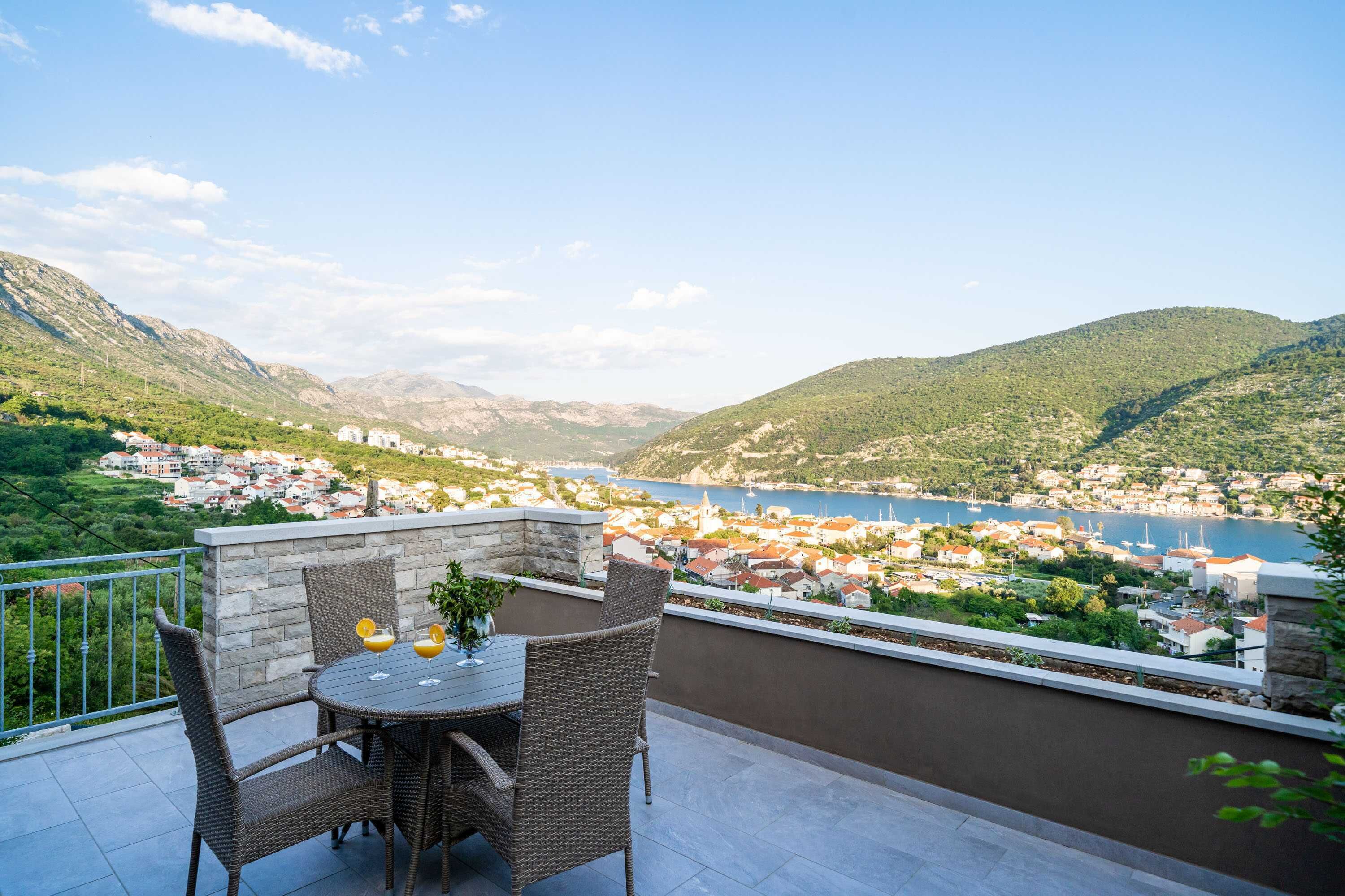 A terrace with wicker furniture and a table, overlooking a town and a lake with mountains in the background.