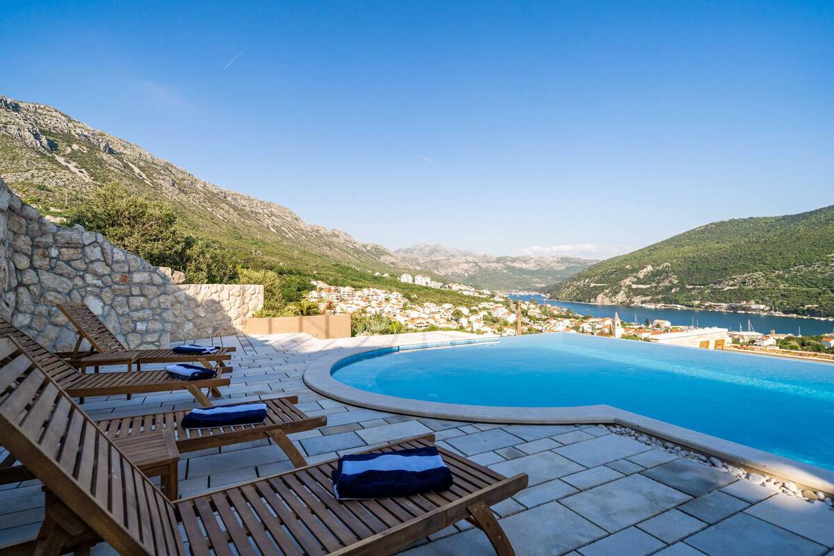 Zona junto a la piscina con tumbonas de madera y pared de piedra, con vistas a un pueblo costero y montañas.