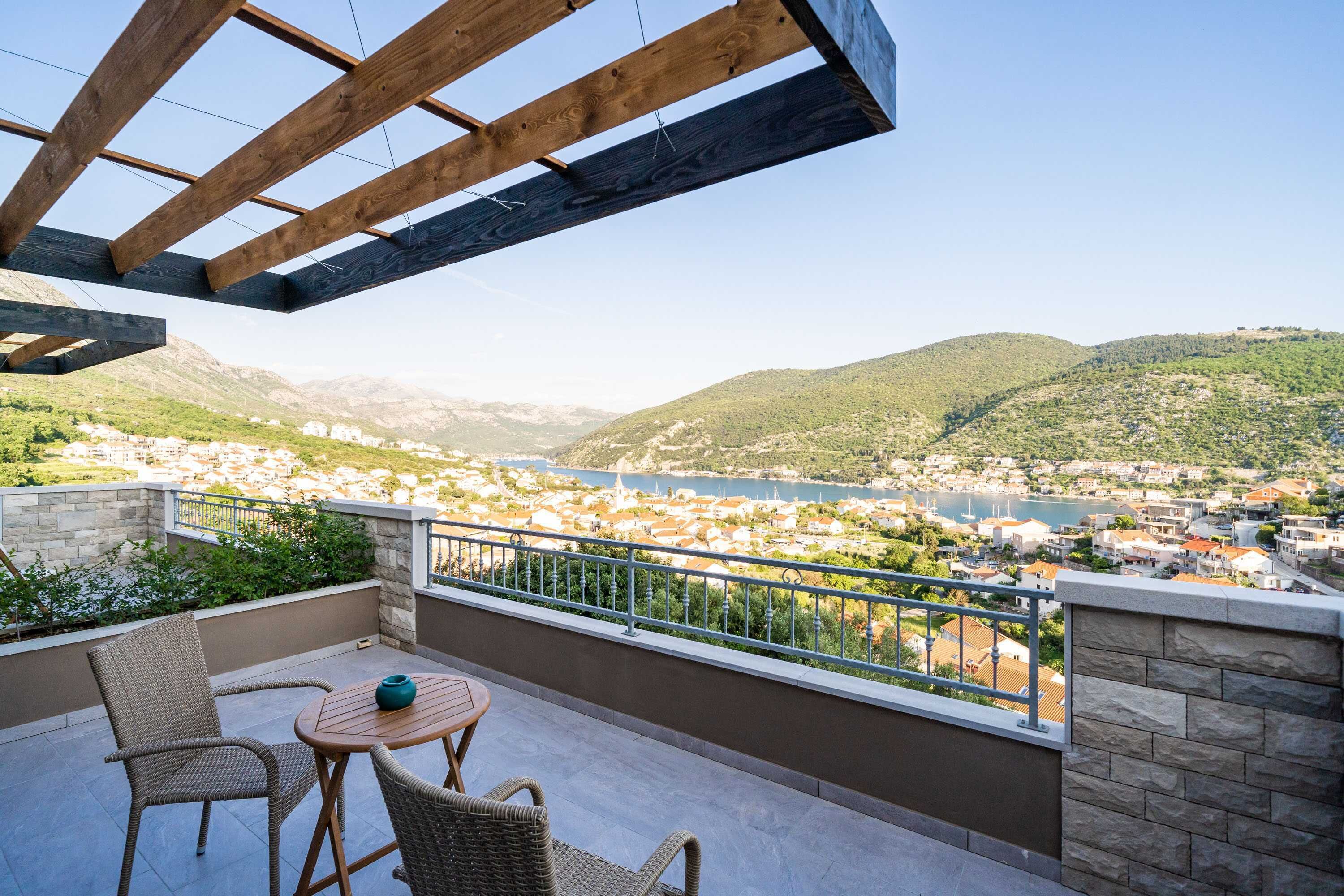 A balcony with wicker chairs and a table, overlooking a town and hills.