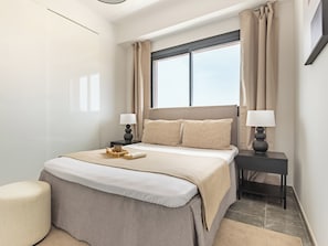 A modern hotel room with a large bed, two bedside tables with lamps, a window with curtains, and a small side table.